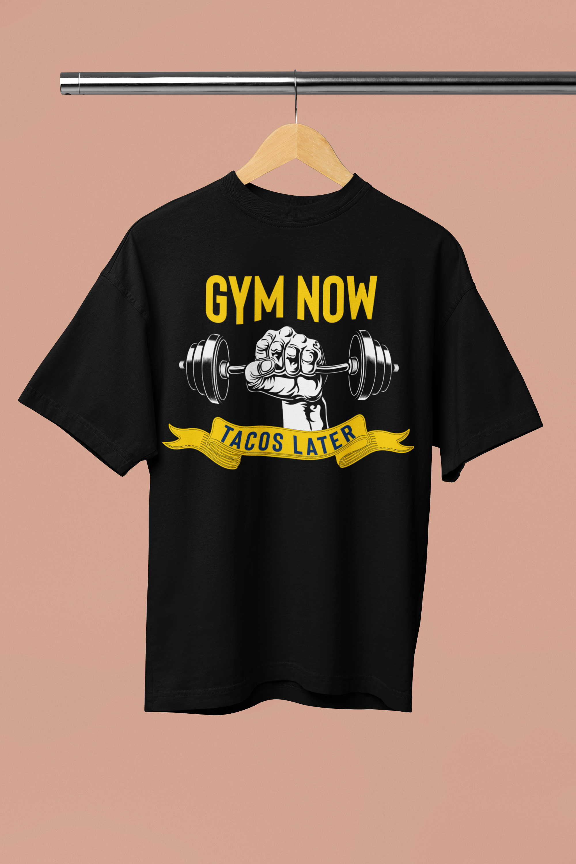 A black oversized unisex t-shirt with the text 'GYM NOW' and 'TACOS LATER' on the front, displayed on a hanger against a pink background.
